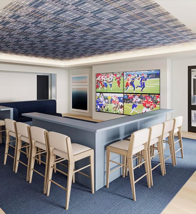 Clubhouse interior featuring large windows, blue plaid wallpaper on the ceiling, light wood flooring, four TVs on the wall, and a combination of couch and bar seating.